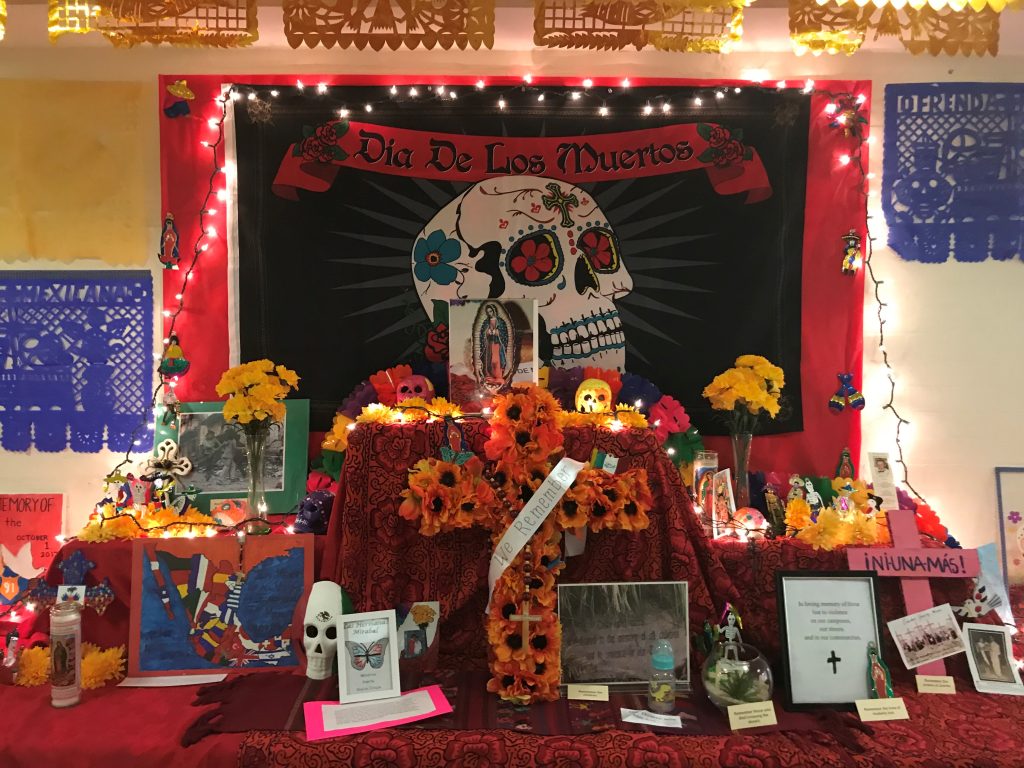 In 2017, a Day of the Dead altar at Notre Dame of Maryland University in Baltimore includes a banner with a colorful skull hanging over bright flowers, Catholic symbols, art and photos of loved ones. (Photo courtesy of Notre Dame of Maryland University) 