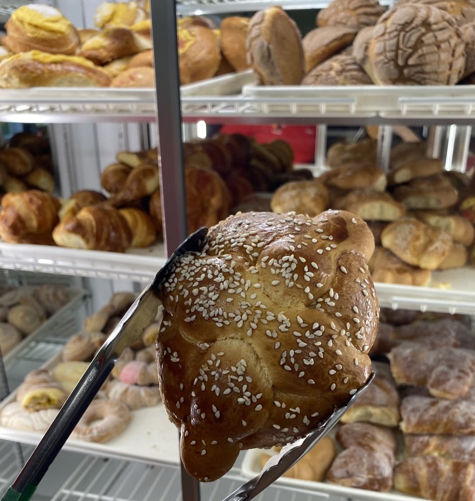 A traditional Mexican pastry called “bread of the dead” — pan de muerto in Spanish — is coated with sesame seeds in a pair of tongs at La Flor de Puebla Bakery in Hyattsville, Maryland, on Oct. 8, 2021. (Photo: Trisha Ahmed / Capital News Service)