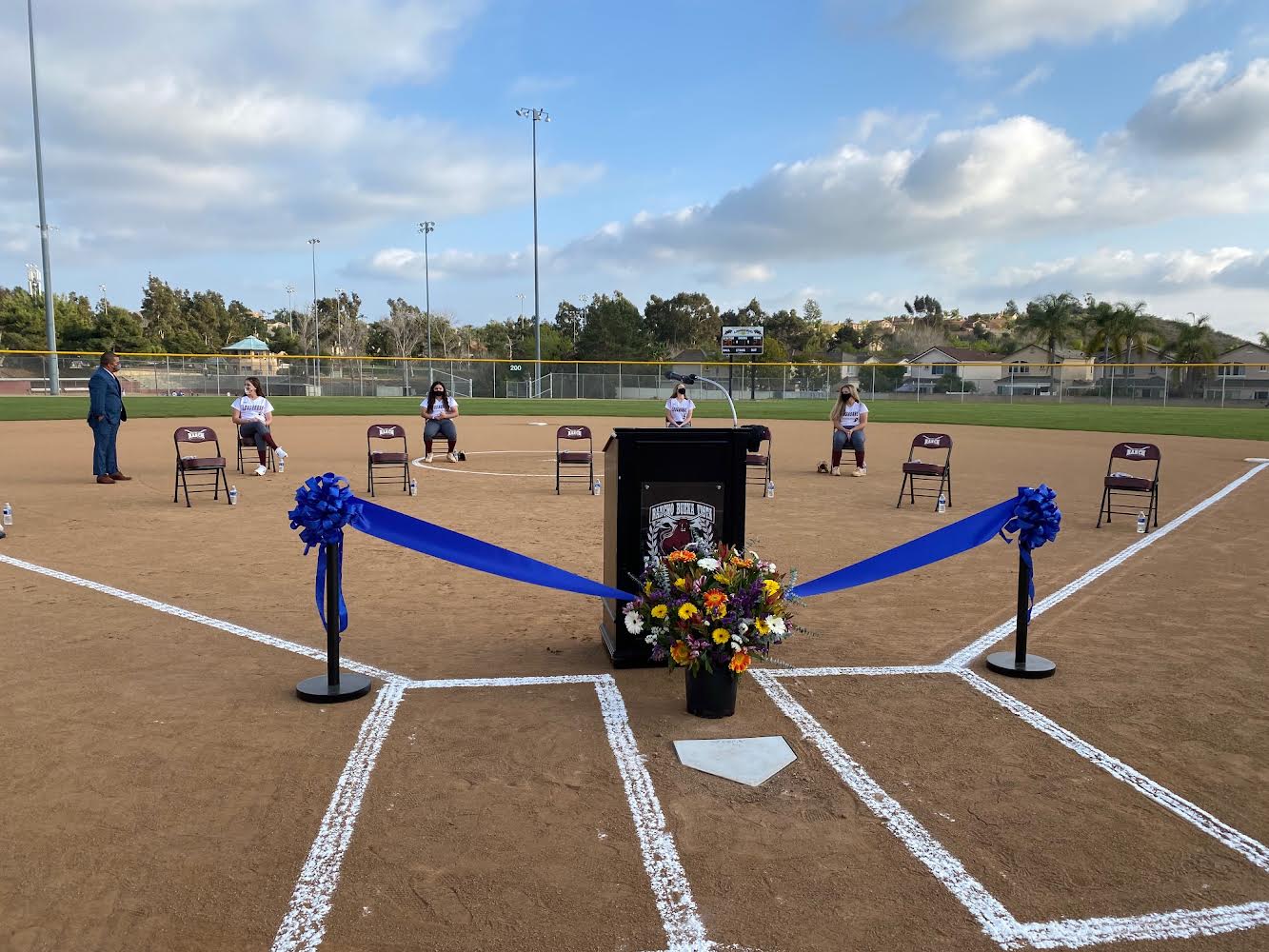 A podium with a ribbon and flowers in front of it with students sitting in the background on the new softball field