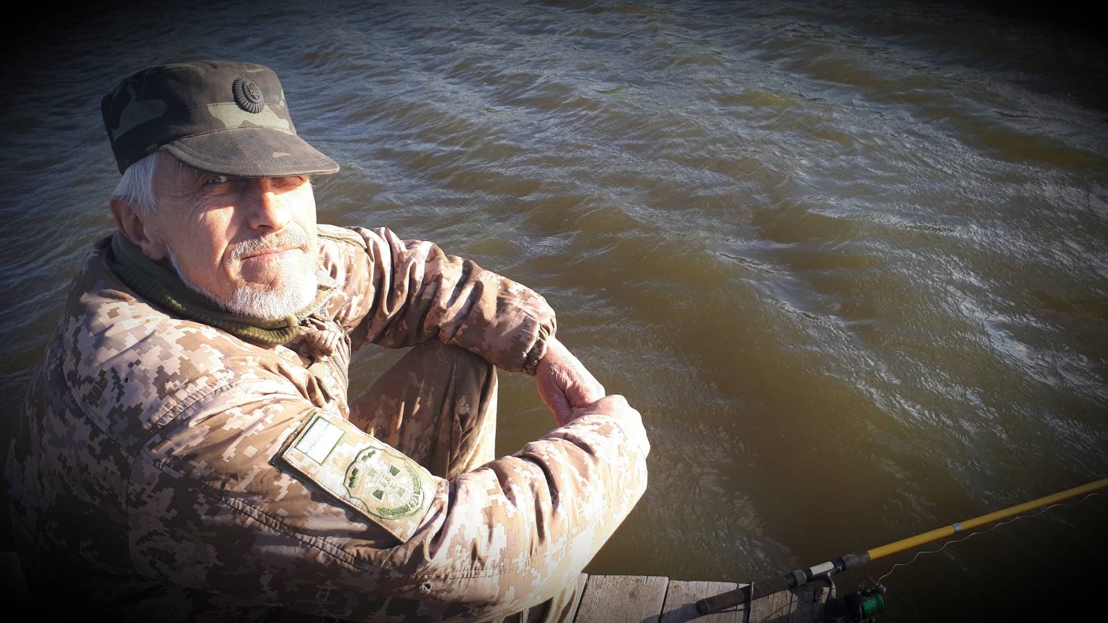 A man in a Ukrainian soldier uniform sits on a dock next to water.