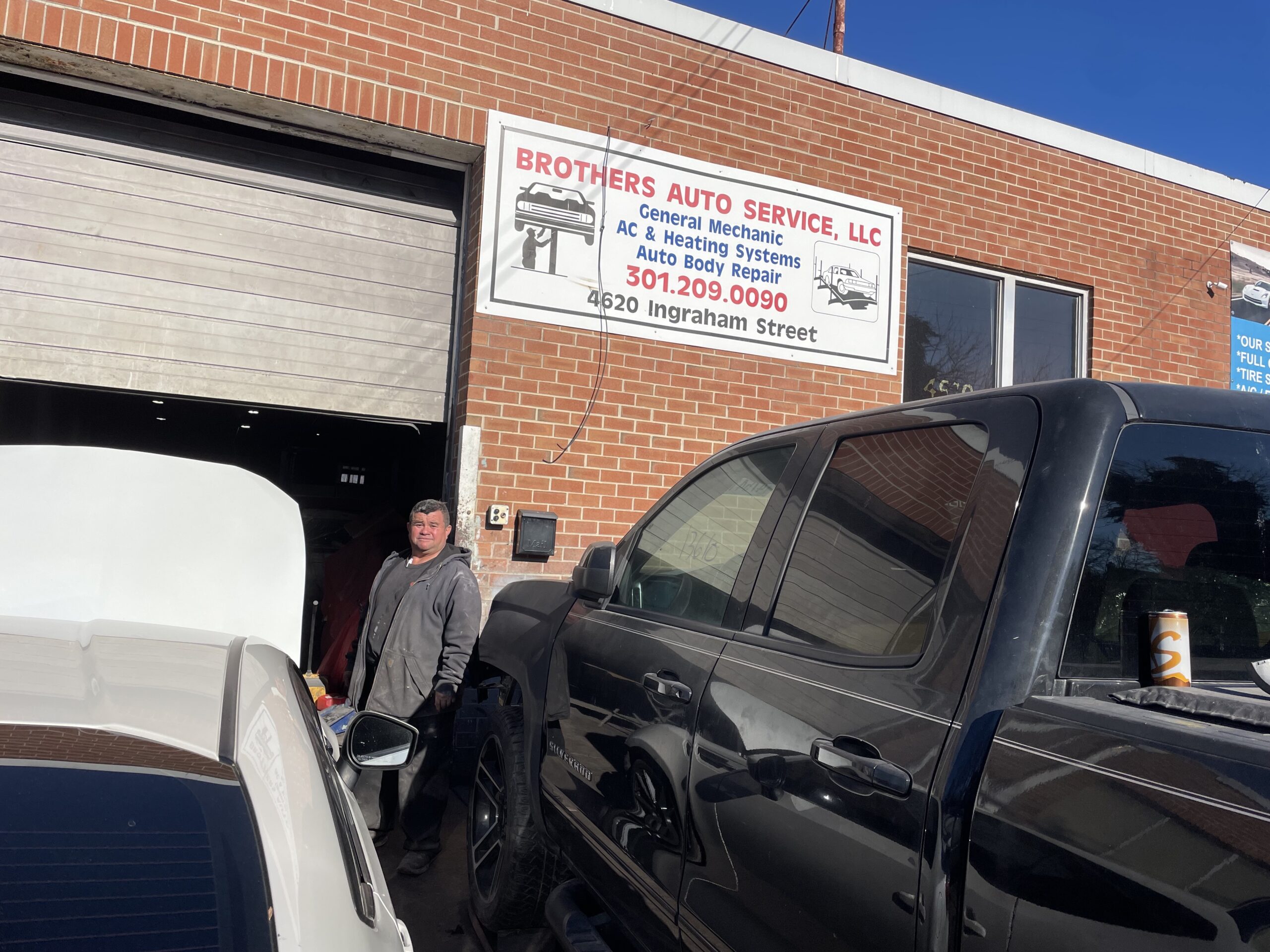 A man stands outside of a an auto shop garage in between a black truck and a white sedan.