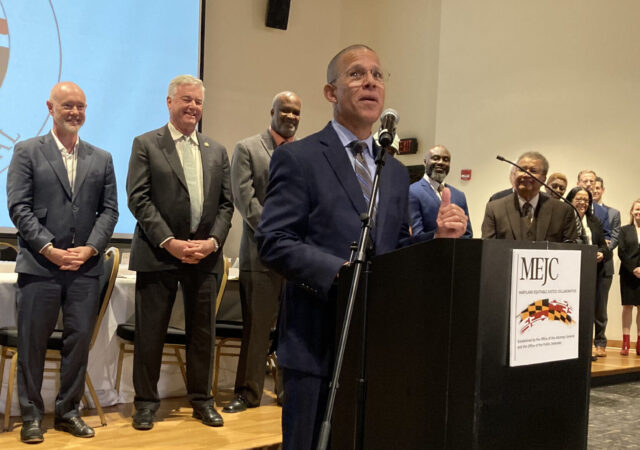 Maryland Attorney General Anthony Brown stands at a podium speaking with a group of lawmakers, agency heads and community organization leaders behind him.
