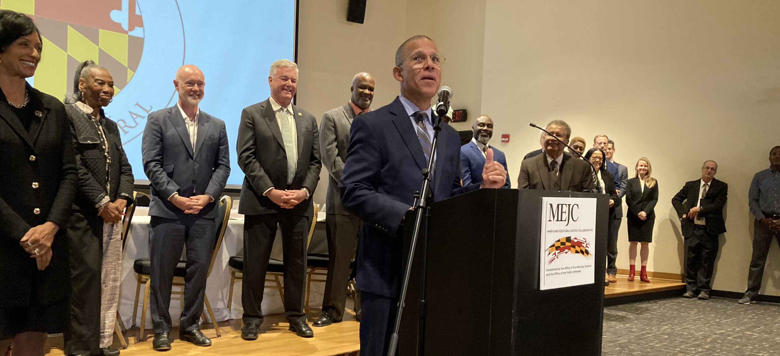 Maryland Attorney General Anthony Brown stands at a podium speaking with a group of lawmakers, agency heads and community organization leaders behind him.