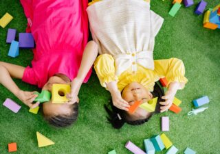 Two young girls lay on a green carpet, playing with colorful blocks.