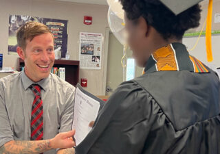 A recent high school graduate wearing a black cap and gown shakes hands with his principal as he receives his diploma.