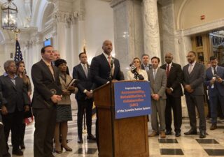 Maryland Gov. Wes Moore speaks from a podium during a press conference in the State House, surrounded by Democratic lawmakers.
