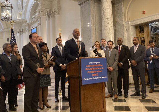 Maryland Gov. Wes Moore speaks from a podium during a press conference in the State House, surrounded by Democratic lawmakers.