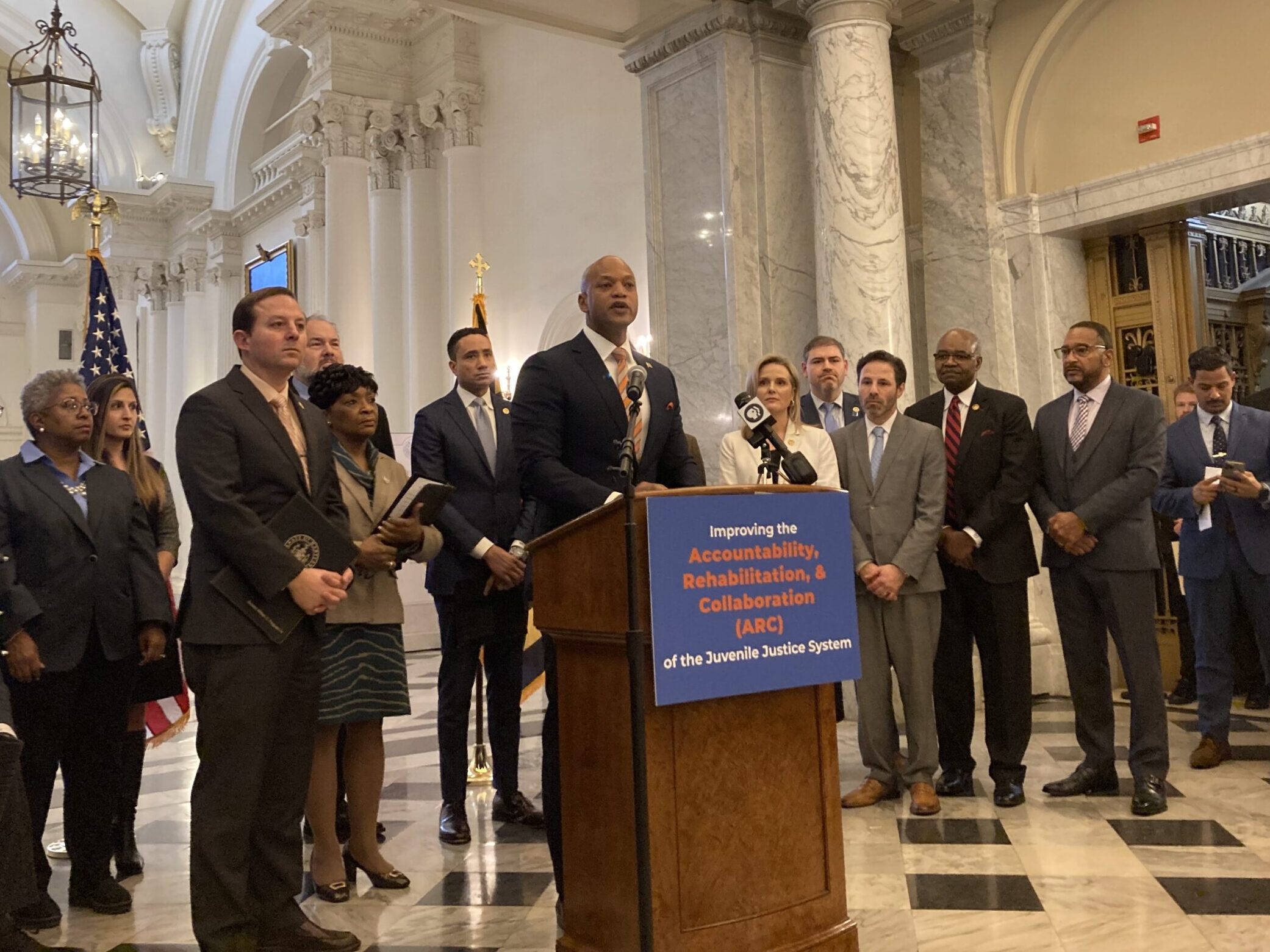 Maryland Gov. Wes Moore speaks from a podium during a press conference in the State House, surrounded by Democratic lawmakers.
