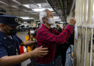 Un perfil lateral de José Rubén Zamora mientras sostiene sus manos esposadas contra los barrotes.