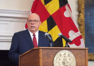 Hogan stands at podium speaking into a microphone with a Maryland flag behind him.