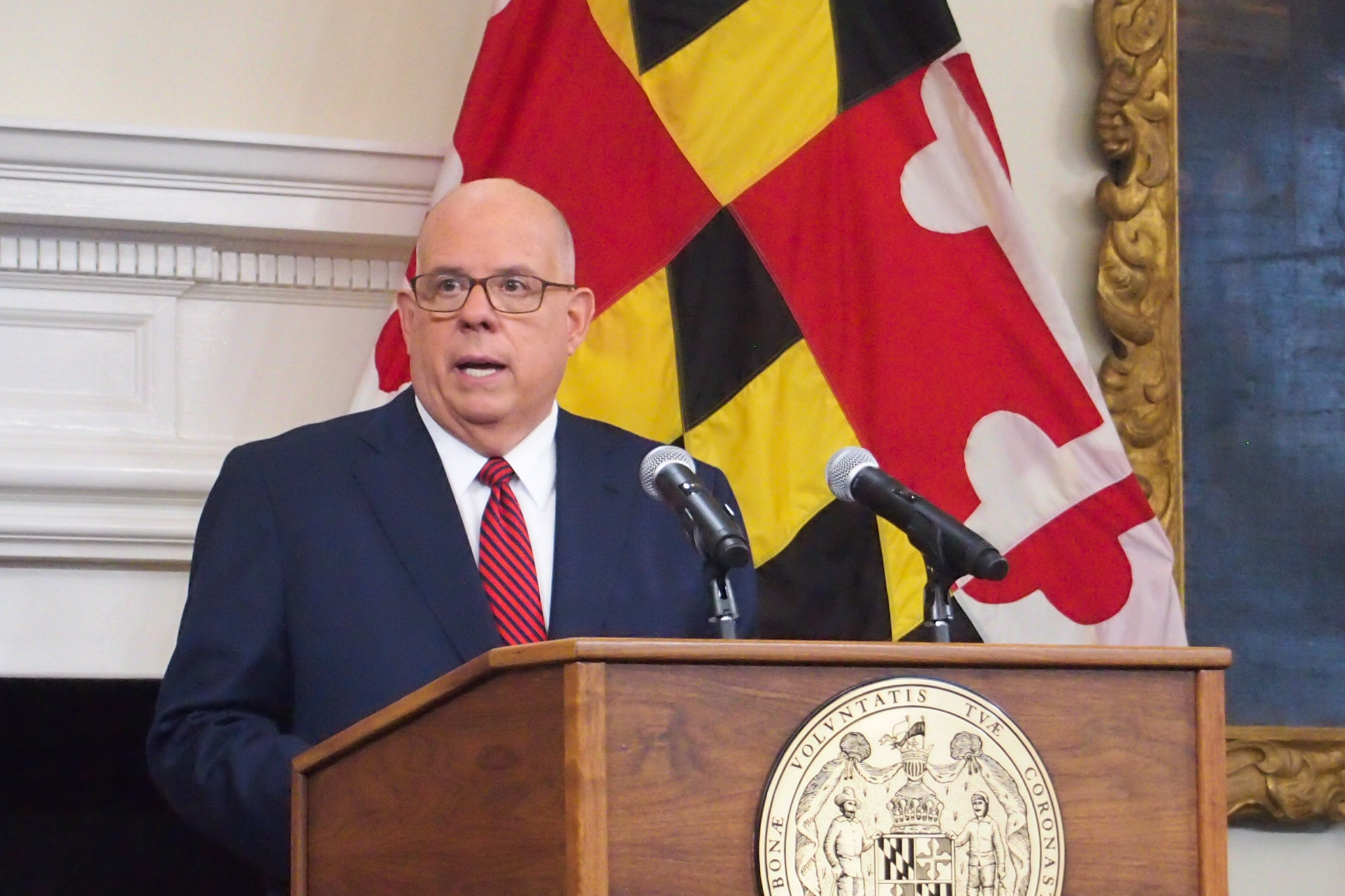 Hogan stands at podium speaking into a microphone with a Maryland flag behind him.