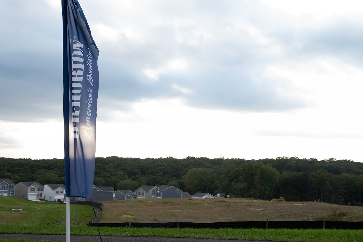 A landscape view of Ridgeley’s Reserve. Homes recently developed are visible on the left-hand side of the site. An open barren field is visible to the right, where more homes will be constructed. A flag bearing the name, “D.R. Horton,” is at the forefront. 