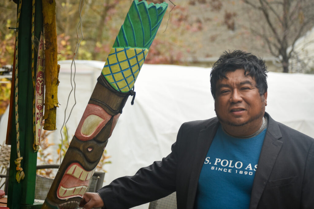 Man in a t-shirt and blazer leans against a wooden pole.