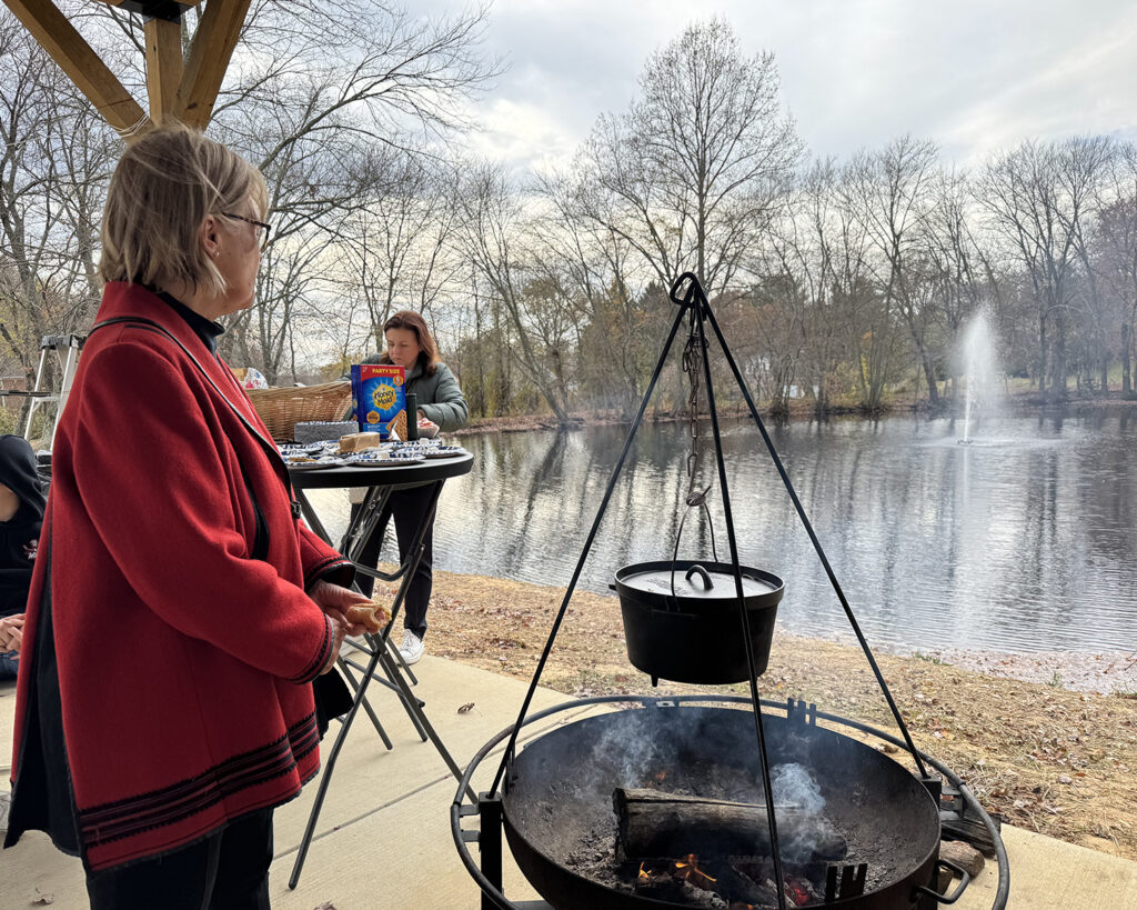 A woman stands near a cauldron beside a body of water. The trees are bare.