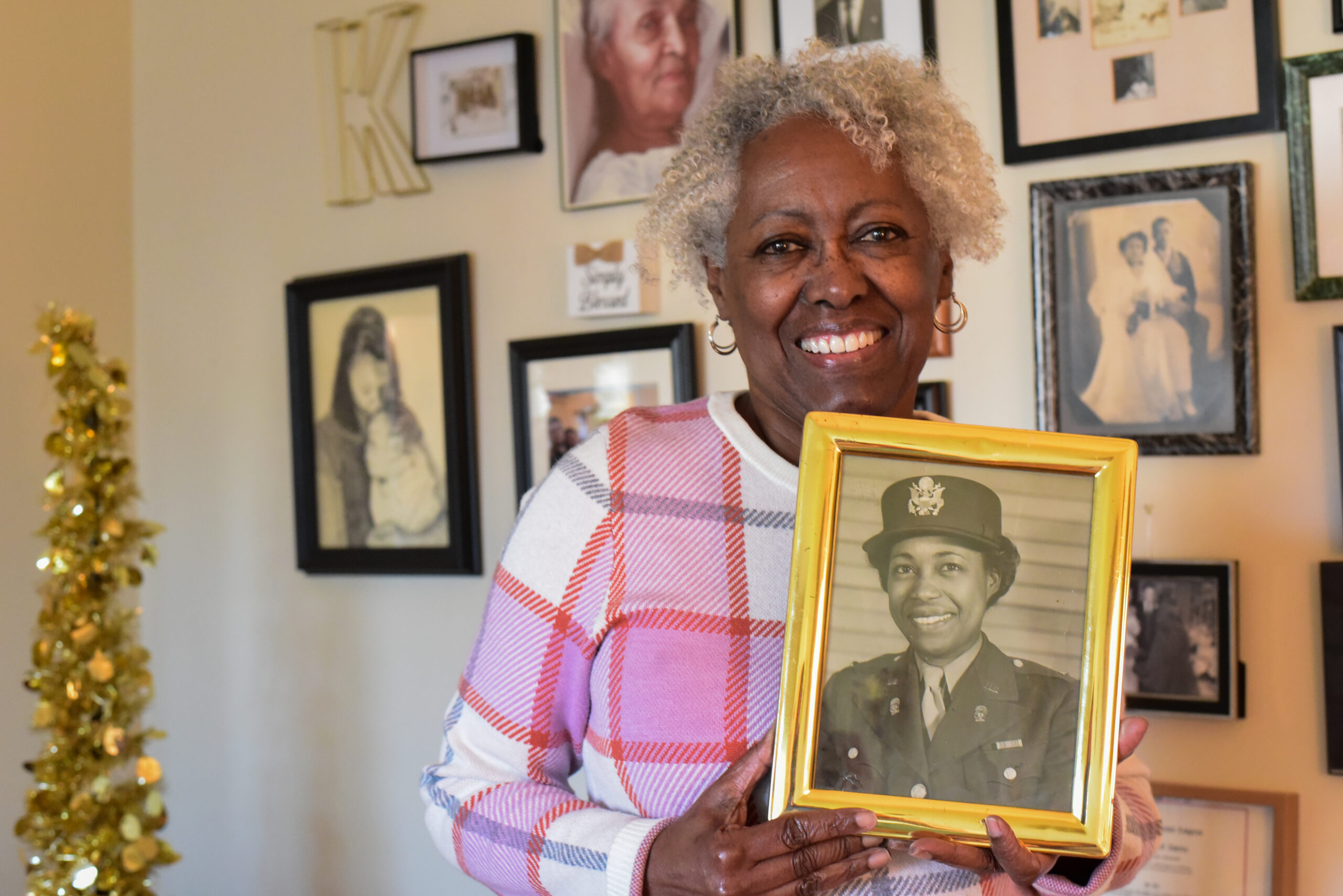 Woman stand in front of a wall of pictures hoisting a picture of her mother.