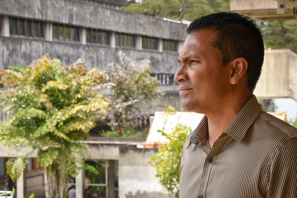 University professor gazes into the distance in front of concrete buildings and trees.